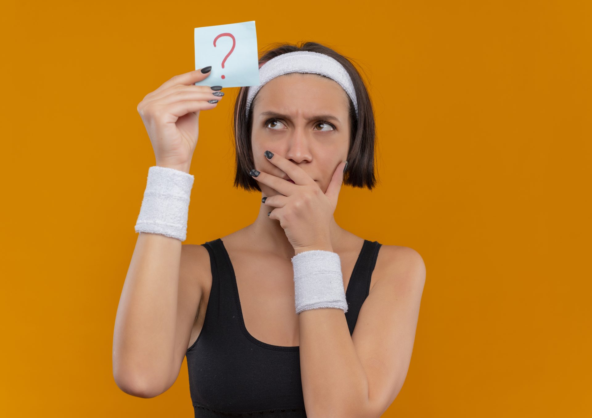 young-fitness-woman-sportswear-with-headband-holding-reminder-paper-with-question-mark-looking-it-with-pensive-expression-thinking-standing-orange-wall